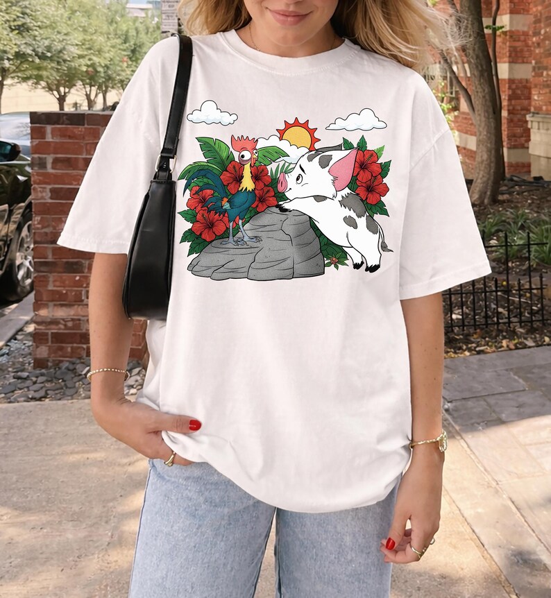 May include: White t-shirt featuring a colorful graphic of a rooster and a pig, surrounded by red flowers, green leaves, clouds, and a sun. The design is set against a light gray rock. The shirt is a casual, oversized style.
