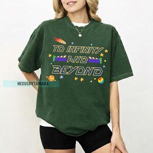 May include: Dark green t-shirt with the phrase "TO INFINITY AND BEYOND" in yellow and blue. The design includes stars, a planet, and a rocket. The shirt is a comfortable, casual style.