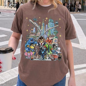 K&ouml;nnte beinhalten: Braunes T-Shirt mit einer farbenfrohen Illustration von Zeichentrickfiguren vor einer Stadtlandschaft mit Hubschraubern und Sternen. Das T-Shirt besteht aus einem weichen Material.