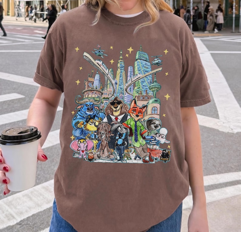 K&ouml;nnte beinhalten: Braunes T-Shirt mit einer farbenfrohen Illustration von Zeichentrickfiguren vor einer Stadtlandschaft mit Hubschraubern und Sternen. Das T-Shirt besteht aus einem weichen Material.
