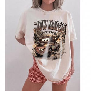 May include: Cream-colored t-shirt featuring a cartoon image of Tow Mater from the movie Cars. The graphic includes the text "Tow Mater" in a distressed font, with the character in action. The shirt is paired with red and white checkered shorts.