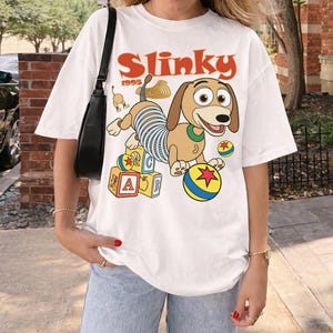 Puede incluir: Camiseta blanca con una imagen de dibujos animados de Slinky Dog de la pel&iacute;cula Toy Story. El dise&ntilde;o incluye la palabra "Slinky" en naranja, el a&ntilde;o "1995" y elementos coloridos como bloques y una pelota.