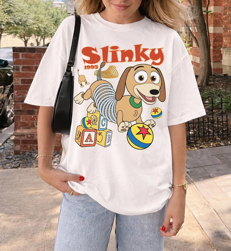 Puede incluir: Camiseta blanca con una imagen de dibujos animados de Slinky Dog de la pel&iacute;cula Toy Story. El dise&ntilde;o incluye la palabra "Slinky" en naranja, el a&ntilde;o "1995" y elementos coloridos como bloques y una pelota.
