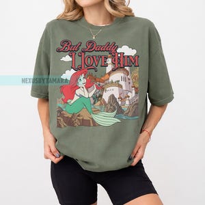 Puede incluir: Camiseta verde oliva con una ilustraci&oacute;n de dibujos animados de una sirena con pelo rojo y cola verde, sentada en una roca. La camiseta tiene el texto "But Daddy I Love Him" en una fuente de estilo vintage, con un castillo en el fondo.