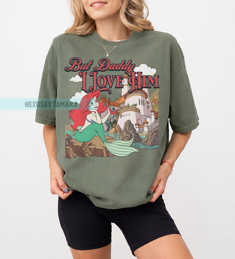 Puede incluir: Camiseta verde oliva con una ilustraci&oacute;n de dibujos animados de una sirena con pelo rojo y cola verde, sentada en una roca. La camiseta tiene el texto "But Daddy I Love Him" en una fuente de estilo vintage, con un castillo en el fondo.