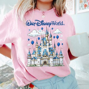 May include: Pink t-shirt featuring a detailed illustration of Cinderella's castle with blue accents, balloons, and clouds. The text "Walt Disney World" is arched above the castle in blue. A white mug is held in the right hand.