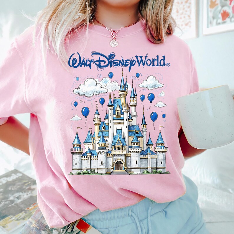May include: Pink t-shirt featuring a detailed illustration of Cinderella's castle with blue accents, balloons, and clouds. The text "Walt Disney World" is arched above the castle in blue. A white mug is held in the right hand.