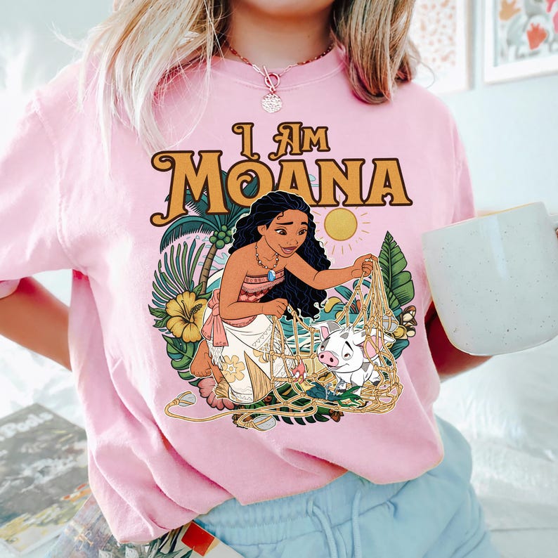 May include: Pink t-shirt featuring a graphic of Moana, a character from the animated film, with the text "I Am Moana." The design includes a sun, palm leaves, flowers, and a pig. The shirt is a crew neck.