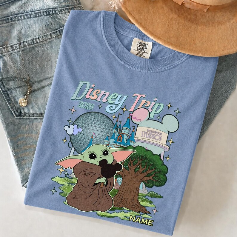 Puede incluir: Camiseta azul claro con un dise&ntilde;o de Baby Yoda sosteniendo una cabeza de Mickey Mouse, con el texto "Disney Trip 2026". El dise&ntilde;o incluye im&aacute;genes de la bola de Epcot, un castillo y un icono de Hollywood Studios.