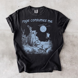 May include: A black short-sleeve t-shirt featuring a light blue graphic print of a frog playing a guitar, a dragonfly, and a moon with a face. The text "rage consumes me" is printed in light blue above the illustration on the front of the shirt.