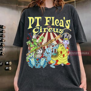 May include: Dark gray t-shirt featuring the text "PT Flea's Circus" in yellow, with a colorful graphic of cartoon insects. The design includes a green grasshopper, blue and yellow caterpillars, and other animated characters.