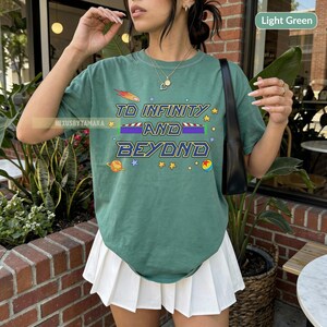 May include: Light green t-shirt with the phrase "TO INFINITY AND BEYOND" in blue and yellow lettering. The design includes a shooting star, planets, and stars. The shirt is paired with a white pleated skirt.