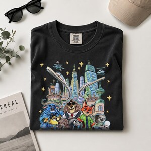 K&ouml;nnte beinhalten: Schwarzes Comfort Colors T-Shirt mit einer farbenfrohen Illustration einer Stadt mit Tierfiguren. Das Design umfasst einen Zug, Hubschrauber und Sterne. Das T-Shirt hat einen Rundhalsausschnitt und ein kleines Etikett.