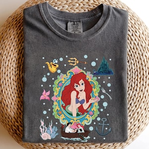 以下が含まれることがあります： ダークグレーのComfort Colors Tシャツ。赤い髪、青い貝殻のブラ、緑の尾を持つ人魚のカラフルなイラストが特徴です。デザインには、城、ボート、その他の海のテーマの要素が含まれています。