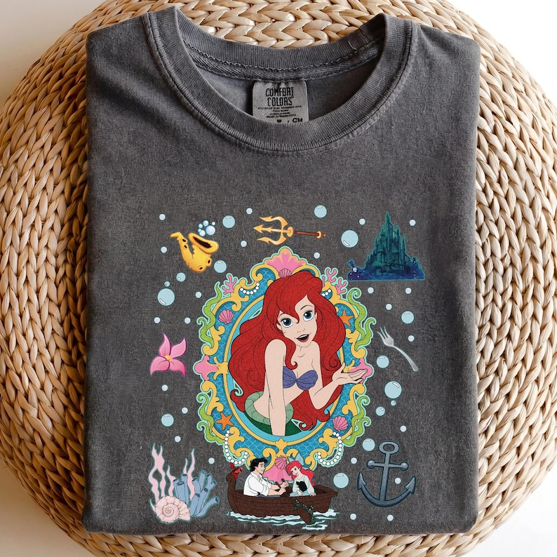 以下が含まれることがあります： ダークグレーのComfort Colors Tシャツ。赤い髪、青い貝殻のブラ、緑の尾を持つ人魚のカラフルなイラストが特徴です。デザインには、城、ボート、その他の海のテーマの要素が含まれています。