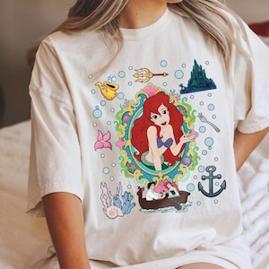 以下が含まれることがあります： クリーム色のTシャツ。赤い髪の毛のマーメイドのカラフルなグラフィックが特徴で、海のテーマのイラストに囲まれています。デザインには三叉の槍、泡、お城が含まれており、気まぐれで芸術的な美学を作り出しています。