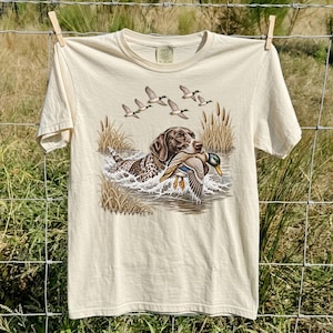 以下が含まれることがあります： クリーム色の半袖Tシャツで、茶色と白の斑点模様の犬が水の中を泳ぎながらマガモを口にくわえているグラフィックプリントが施されており、背景には4羽の飛んでいるカモと背の高い葦が描かれています。