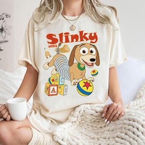 Puede incluir: Camiseta color crema con una ilustraci&oacute;n de dibujos animados de Slinky Dog de la pel&iacute;cula Toy Story. El dise&ntilde;o incluye la palabra "Slinky" en rojo, el a&ntilde;o "1995" y bloques y pelotas de juguete coloridos.