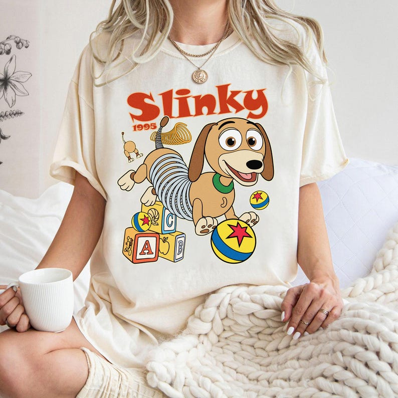 Puede incluir: Camiseta color crema con una ilustraci&oacute;n de dibujos animados de Slinky Dog de la pel&iacute;cula Toy Story. El dise&ntilde;o incluye la palabra "Slinky" en rojo, el a&ntilde;o "1995" y bloques y pelotas de juguete coloridos.