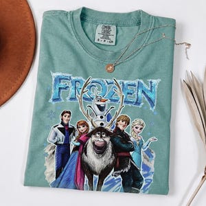 May include: A light teal Comfort Colors t-shirt with the word "FROZEN" in a blue, icy font, and cartoon characters from the film. The shirt has a crew neck and short sleeves. A gold necklace with a pendant is on the shirt.