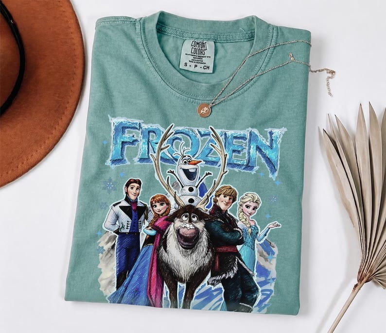 May include: A light teal Comfort Colors t-shirt with the word "FROZEN" in a blue, icy font, and cartoon characters from the film. The shirt has a crew neck and short sleeves. A gold necklace with a pendant is on the shirt.