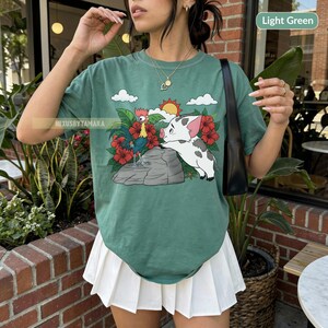 May include: Light green t-shirt featuring a cartoon illustration of a rooster and a pig, with red flowers, clouds, and a sun. The text "NexusbyTamara" is visible on the shirt. The shirt is paired with a white pleated skirt.