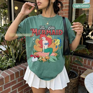 May include: Light green t-shirt with a graphic of Ariel from The Little Mermaid. The design shows Ariel on a seashell, with Flounder, Sebastian, and other sea creatures. The text "The Little Mermaid" is above the graphic.