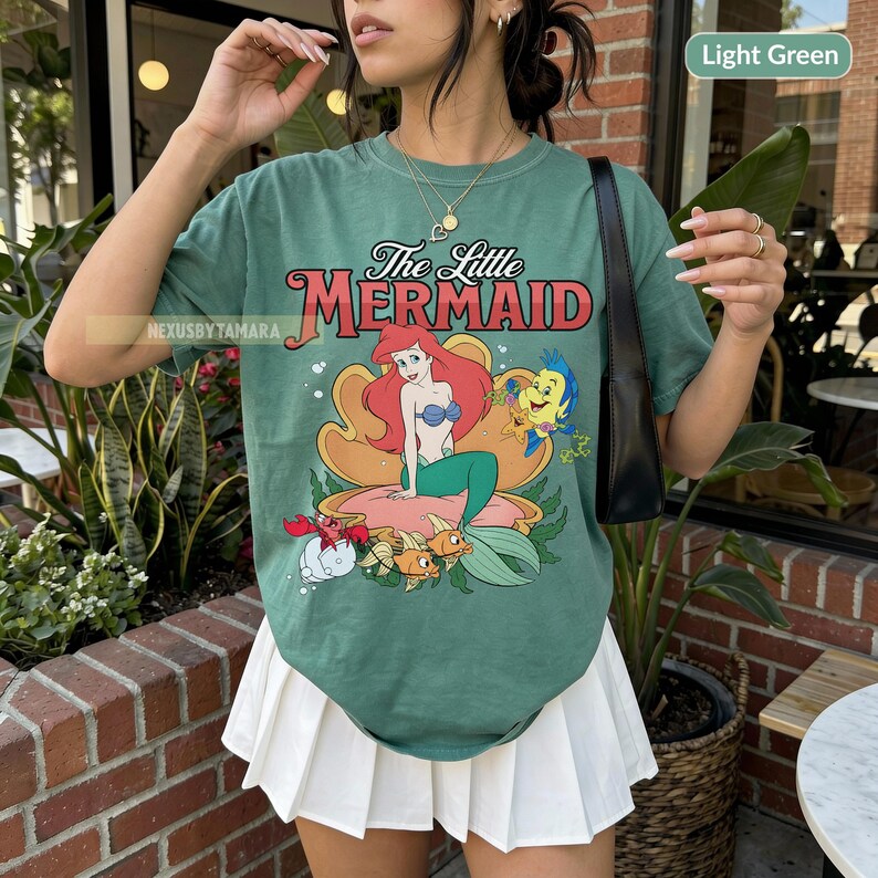 May include: Light green t-shirt with a graphic of Ariel from The Little Mermaid. The design shows Ariel on a seashell, with Flounder, Sebastian, and other sea creatures. The text "The Little Mermaid" is above the graphic.
