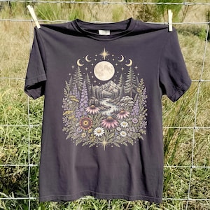 Op de afbeelding: Een antracietgrijs T-shirt met korte mouwen en een centrale grafische print van een volle maan, sterrenhemel, bergketen en een stromende rivier, omringd door een kleurrijk arrangement van wilde bloemen, waaronder paarse lupinen en madeliefjes.