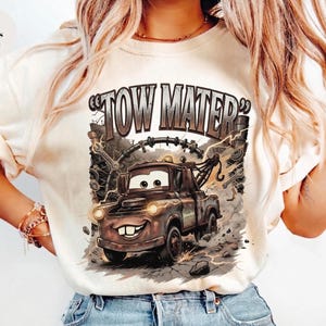 May include: Ivory-colored t-shirt featuring a graphic of Tow Mater from the Cars movie. The design includes the text "TOW MATER" in a distressed font, with the character's image and background details.
