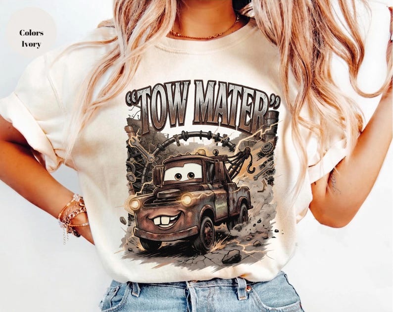 May include: Ivory-colored t-shirt featuring a graphic of Tow Mater from the Cars movie. The design includes the text "TOW MATER" in a distressed font, with the character's image and background details.