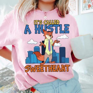 以下が含まれることがあります： 「It's Called A Hustle Sweetheart」の文字と漫画のキツネのキャラクターが描かれたピンク色のTシャツ。キツネはシャツとネクタイを着ており、背景には街並みが描かれています。Tシャツは明るいピンク色です。