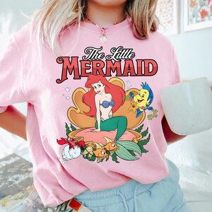 May include: Pale pink t-shirt with a graphic of Ariel from The Little Mermaid. The design includes Ariel with red hair, a blue seashell bra, and a green tail, along with Flounder and Sebastian. The text "The Little Mermaid" is above the graphic.