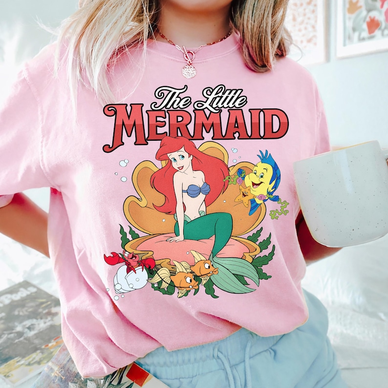 May include: Pale pink t-shirt with a graphic of Ariel from The Little Mermaid. The design includes Ariel with red hair, a blue seashell bra, and a green tail, along with Flounder and Sebastian. The text "The Little Mermaid" is above the graphic.