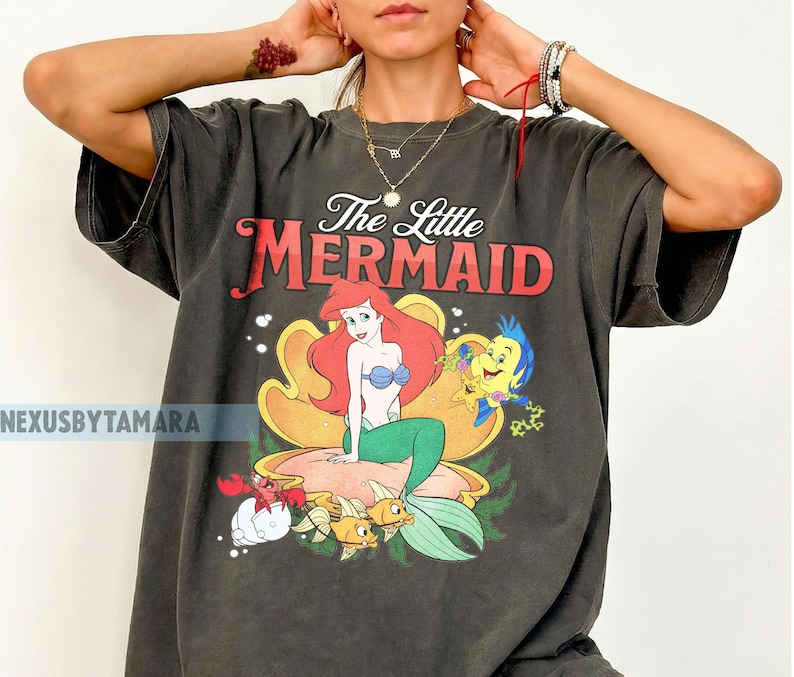 May include: Dark grey t-shirt with a graphic of Ariel from The Little Mermaid. The design features Ariel with red hair, a blue top, and green tail, alongside Flounder, Sebastian, and other sea creatures. The text "The Little Mermaid" is above the graphic.