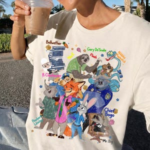 Op de afbeelding: Cr&egrave;mekleurig T-shirt met een kleurrijke cartoonillustratie van verschillende dierenpersonages. Het ontwerp bevat personages zoals een luiaard, een vos en een konijn, met tekstlabels zoals "Judy" en "Chief Bogo".
