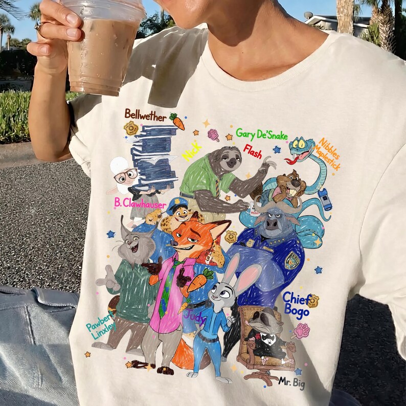 Op de afbeelding: Cr&egrave;mekleurig T-shirt met een kleurrijke cartoonillustratie van verschillende dierenpersonages. Het ontwerp bevat personages zoals een luiaard, een vos en een konijn, met tekstlabels zoals "Judy" en "Chief Bogo".