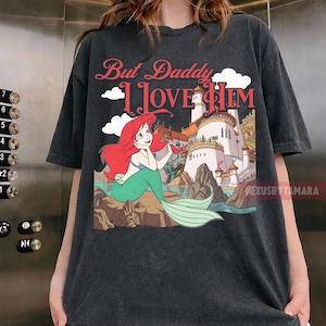 Puede incluir: Camiseta gris oscuro con una sirena de dibujos animados con pelo rojo y cola verde, sentada en una roca. La camiseta tiene el texto "But Daddy I Love Him" en letras rojas sobre la sirena y una ilustraci&oacute;n de castillo.