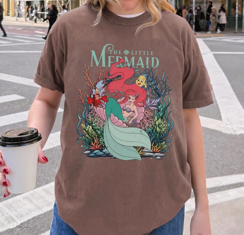 May include: Brown t-shirt with a graphic of Ariel from The Little Mermaid. The design shows Ariel, Flounder, Sebastian, and other characters in an underwater scene. The shirt has a crew neck and short sleeves.