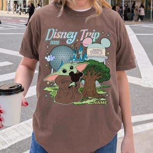 Puede incluir: Camiseta marr&oacute;n con un gr&aacute;fico colorido de Disney Trip 2026 que presenta a Baby Yoda sosteniendo un peluche de Mickey Mouse, un &aacute;rbol e iconos del parque. La camiseta tiene cuello redondo y mangas cortas.