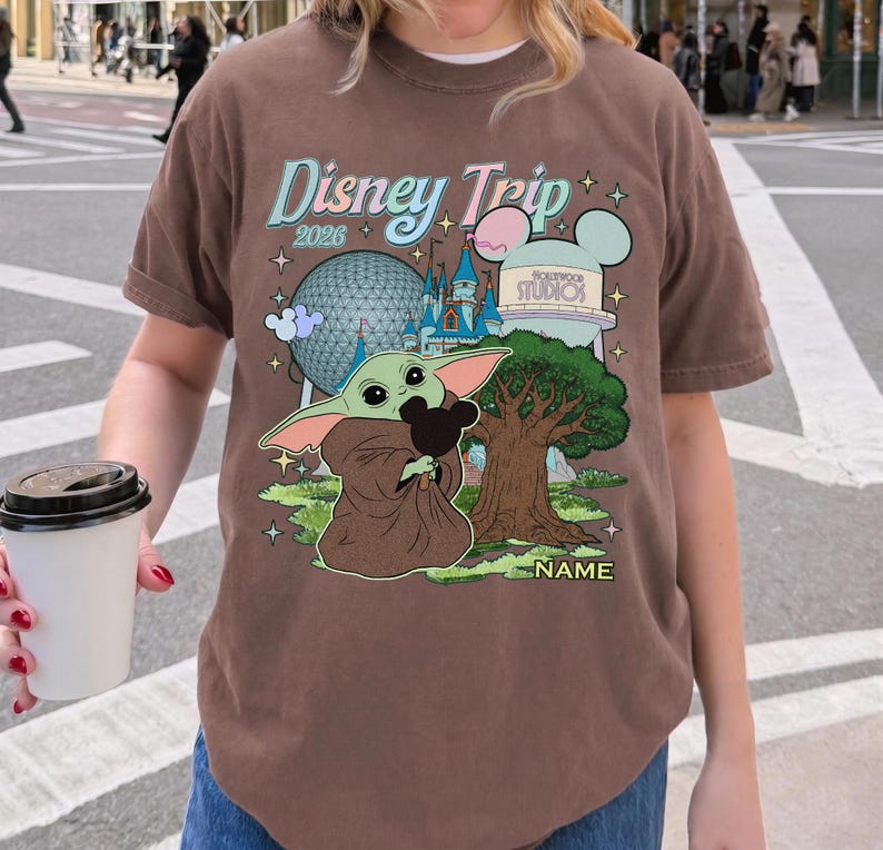 Puede incluir: Camiseta marr&oacute;n con un gr&aacute;fico colorido de Disney Trip 2026 que presenta a Baby Yoda sosteniendo un peluche de Mickey Mouse, un &aacute;rbol e iconos del parque. La camiseta tiene cuello redondo y mangas cortas.