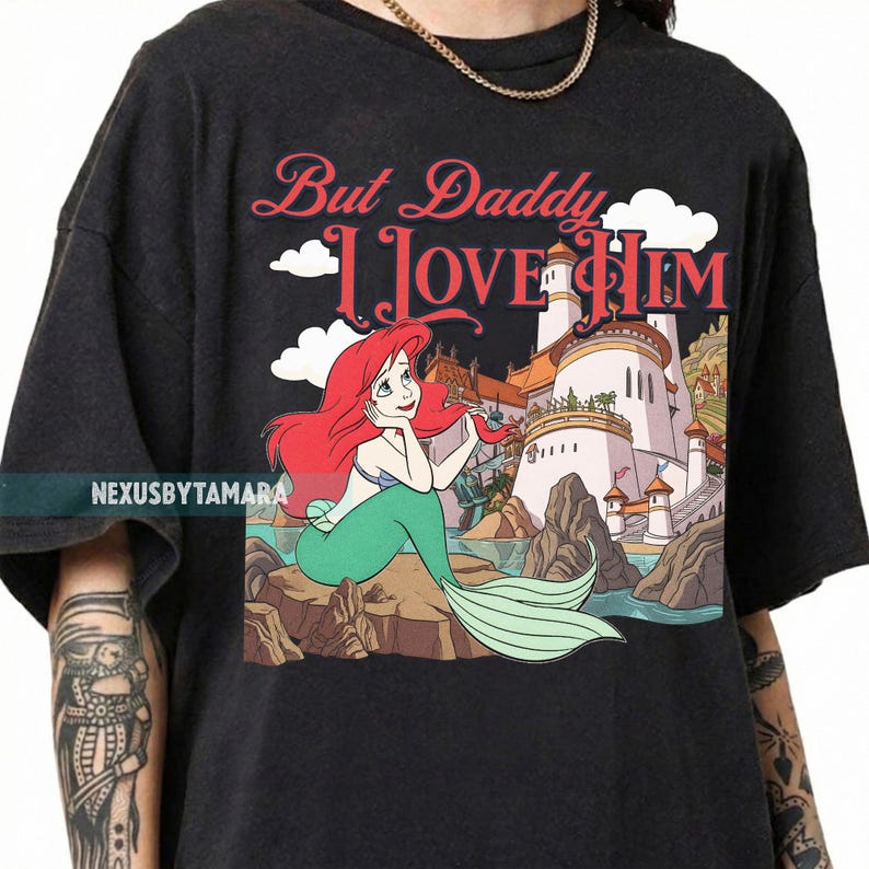 Puede incluir: Camiseta gris oscuro con una ilustraci&oacute;n de dibujos animados de una sirena con cabello rojo y cola verde. El texto "But Daddy I Love Him" est&aacute; encima de la sirena, con un castillo al fondo.