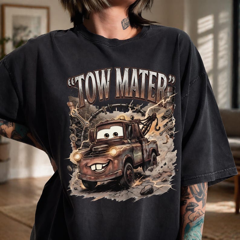 May include: Black t-shirt featuring a graphic of Tow Mater from the movie Cars. The design includes the text "TOW MATER" in a distressed font, with the character's image in a detailed, cartoon style. The shirt has a vintage, worn look.