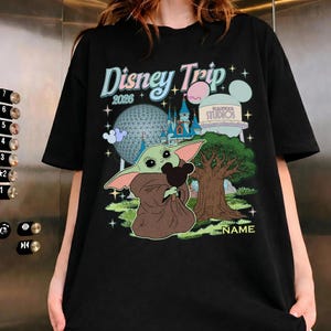 Puede incluir: Camiseta negra con un colorido gr&aacute;fico de Disney Trip 2026. El dise&ntilde;o presenta a Baby Yoda sosteniendo una cabeza de Mickey Mouse, un castillo, un &aacute;rbol y la torre de agua de Hollywood Studios. La camiseta tiene una secci&oacute;n de nombre personalizada.