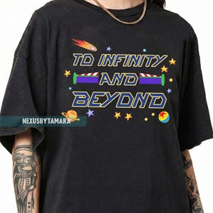 May include: Black t-shirt with the phrase "TO INFINITY AND BEYOND" in a colorful, cartoonish font. The design includes stars, a comet, and a planet. The shirt has a relaxed fit and short sleeves.