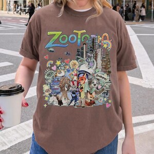 May include: Brown t-shirt featuring the word "Zootopia" in colorful lettering above a detailed graphic of characters and cityscapes from the animated film. The design includes Judy Hopps, Nick Wilde, and other characters.