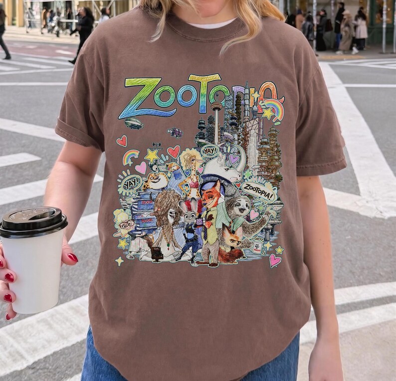 May include: Brown t-shirt featuring the word "Zootopia" in colorful lettering above a detailed graphic of characters and cityscapes from the animated film. The design includes Judy Hopps, Nick Wilde, and other characters.