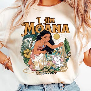 May include: Cream-colored t-shirt featuring the text "I Am Moana" in a retro font. The graphic depicts Moana with a pig, surrounded by tropical foliage and a sun. The design is colorful and detailed.