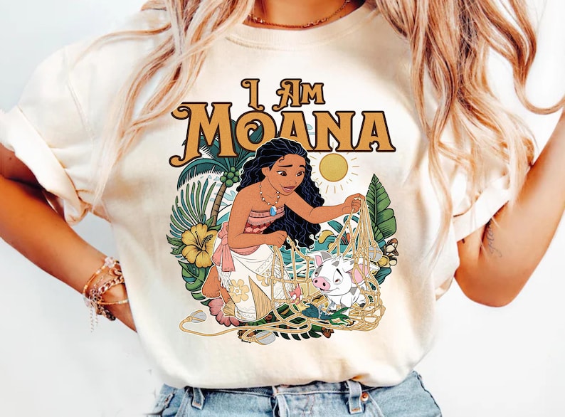 May include: Cream-colored t-shirt featuring the text "I Am Moana" in a retro font. The graphic depicts Moana with a pig, surrounded by tropical foliage and a sun. The design is colorful and detailed.