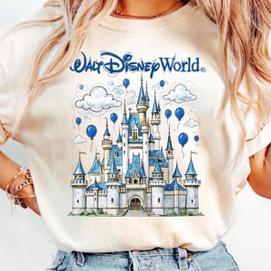 May include: Cream-colored t-shirt featuring a detailed illustration of Cinderella's castle with blue accents, clouds, and balloons. The text "Walt Disney World" is arched above the castle. A whimsical design for Disney fans.
